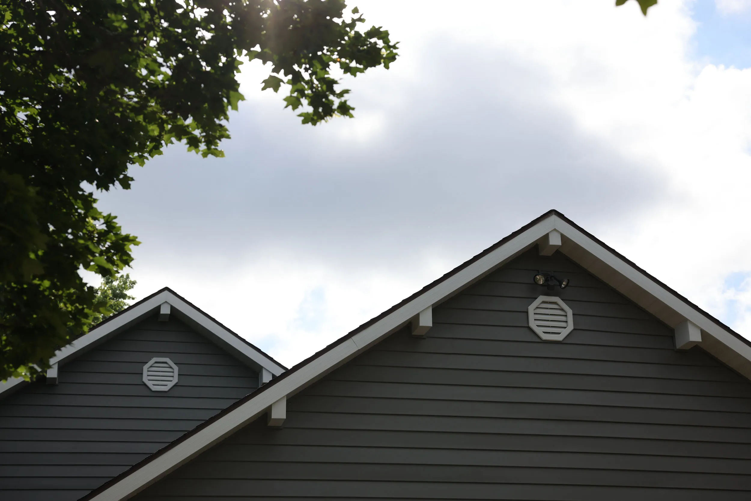 Two gray house gables with white trim and octagonal vents under a cloudy sky with tree leaves framing the top corners.